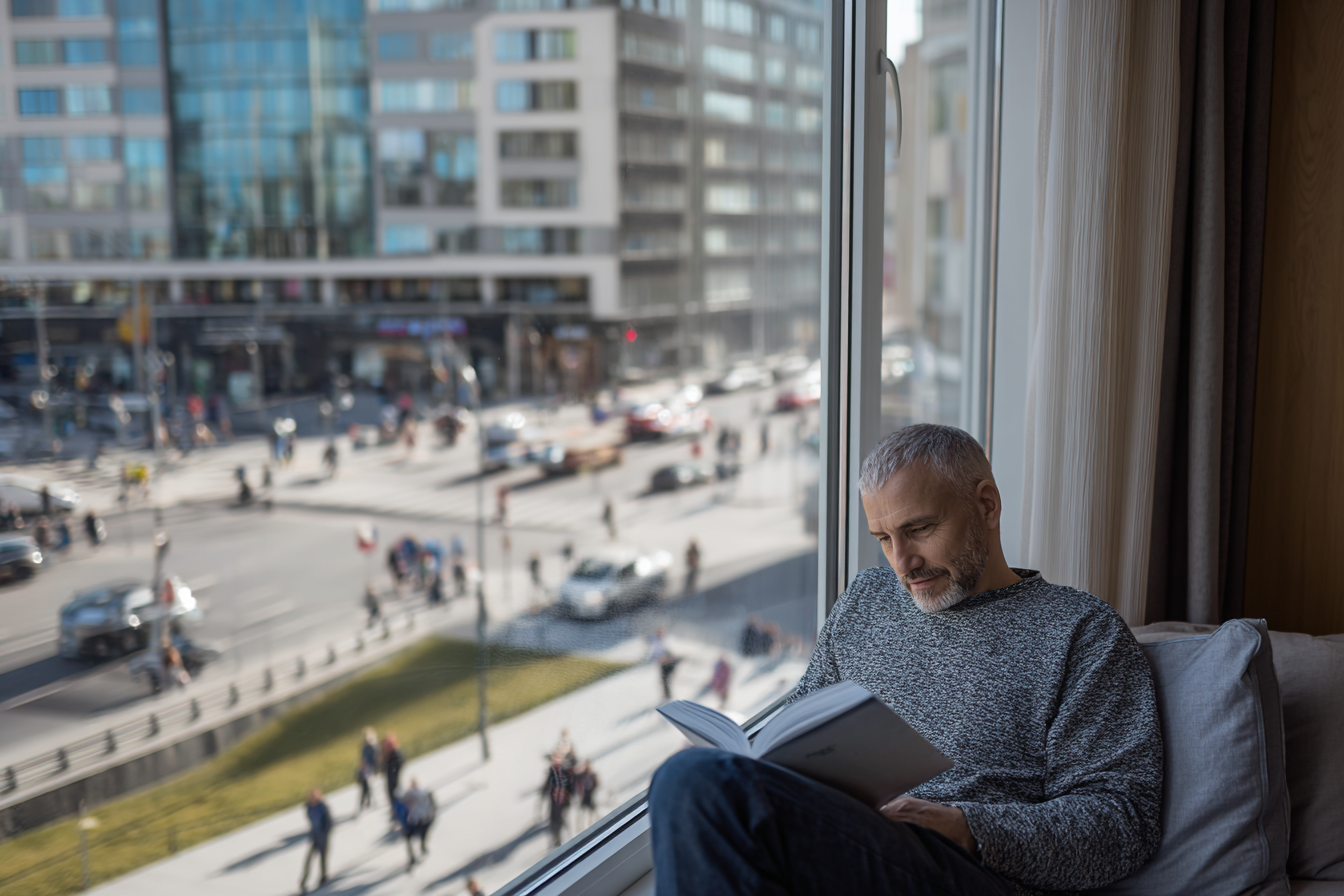 Une personne lit paisiblement près d’une fenêtre en PVC, tandis que l’agitation urbaine reste visible à l’extérieur.